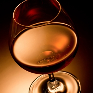 A close crop shows a rounded stemmed glass against a nearly black background. Warm amber and copper highlights curve across the bowl and stem, with a dark red reflection near the rim. No text or other objects are visible.