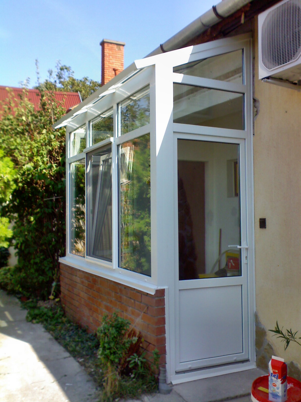 A tall white-framed glazed enclosure is shown from a close, oblique angle beside a cream-colored wall. Its front section includes a hinged door with a white handle, while window panels and a sloping glazed roof continue along the left side. A red brick base and a brick chimney are visible against trees and blue sky. A wall-mounted outdoor air-conditioning unit appears high on the right.