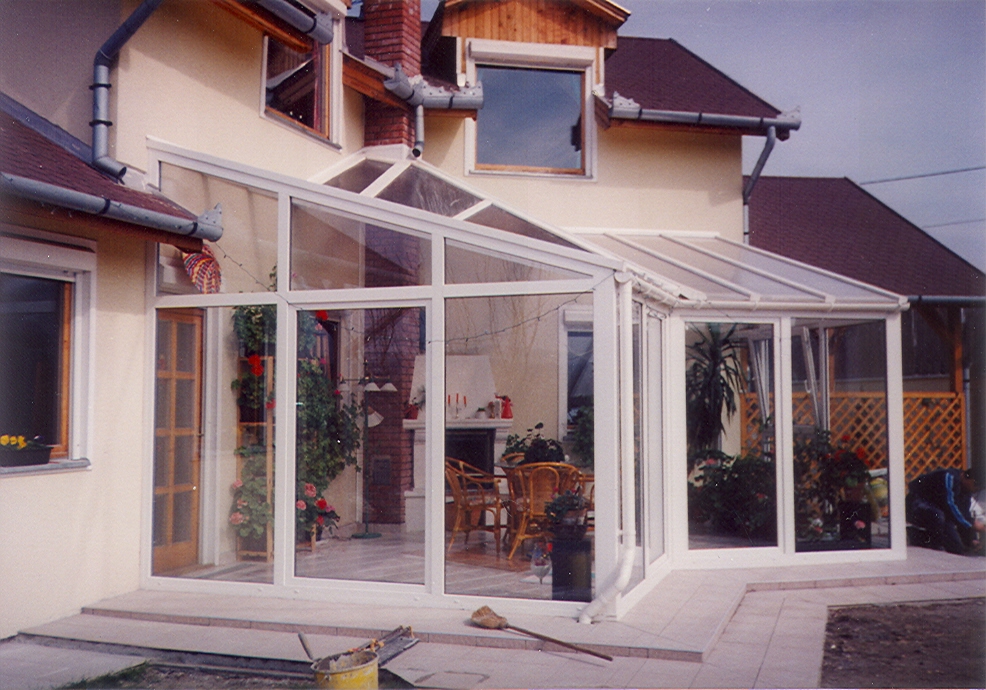 A white-framed glass enclosure stretches across the front of a beige house, with adjoining roof sections sloping toward the patio. The glazing reveals a table, chairs, plants, and other furnishings inside. A paved area with scattered tools or materials fills the foreground, and neighboring rooflines are visible behind the house.