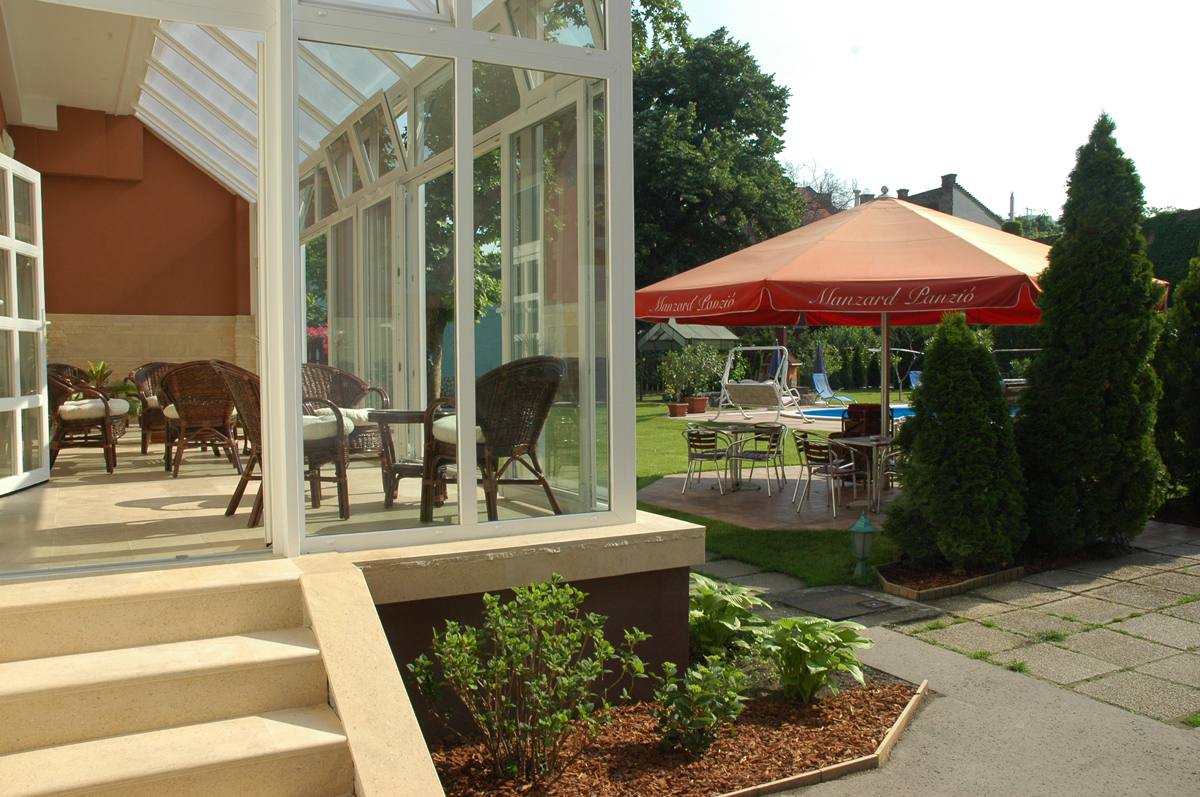 An enclosed glass veranda opens onto a paved garden seating area, with wicker chairs and small tables visible inside. Outside, metal chairs and tables sit beneath a large beige umbrella with the readable words “Manzard Panzió” on its maroon valance. Steps descend from the veranda, and clipped shrubs, trees, and lawn fill the right side. White framing and large panes dominate the left foreground.