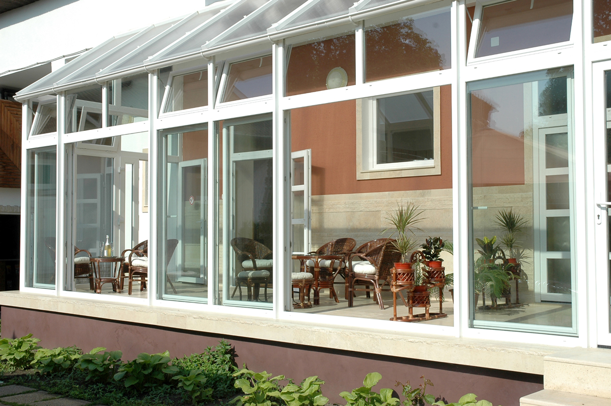 A long white-framed glass veranda runs across the front of a house, with several upper window panels tilted open. Wicker chairs, a small table, and potted plants are visible through the large panes. The interior wall behind the glazing is terracotta above a pale stone band. A narrow garden bed with leafy plants borders the pale ledge at the base.