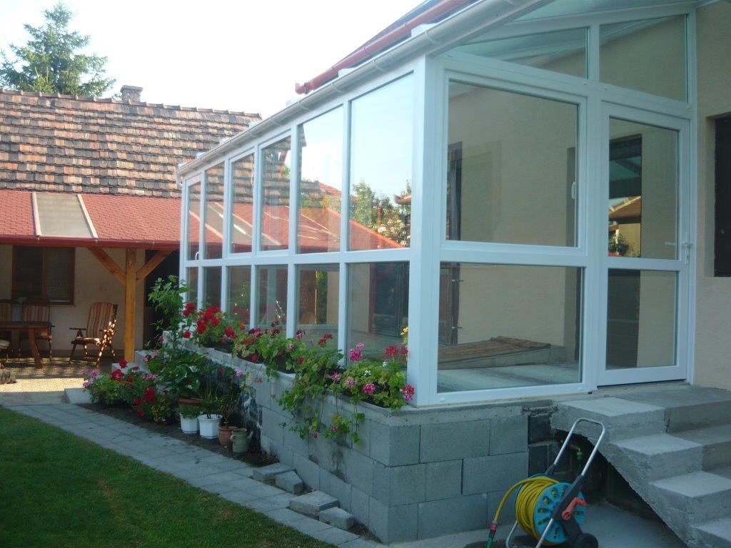 A white-framed glazed conservatory projects from the side of a house, with roof glazing and a long row of windows. Pink and red flowers line the raised edge beneath the glass, while a paved path and patch of lawn occupy the foreground. A covered wooden seating area and tiled roof are visible behind it at left. Bright sunlight creates reflections across the conservatory panes.
