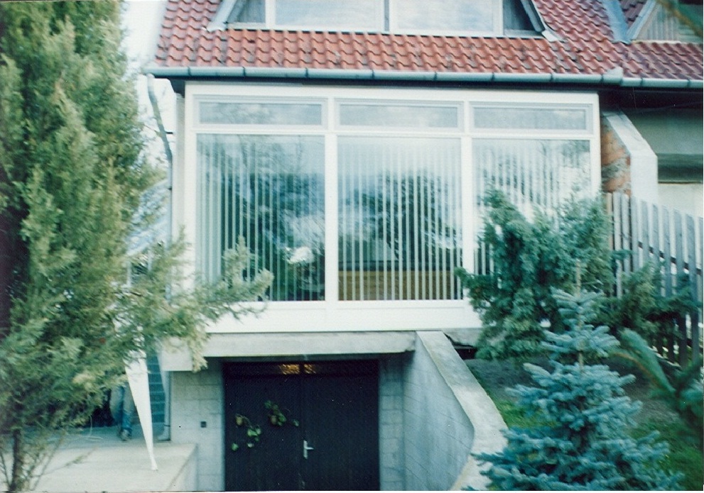 A white-framed glazed enclosure spans the front of a house beneath a red tiled roof. Vertical blinds cover much of the enclosure, and the glass reflects trees and garden greenery. The structure stands above a dark rectangular opening in a gray masonry base, with concrete side walls and steps leading toward the entrance. Trees and shrubs frame both sides of the view.