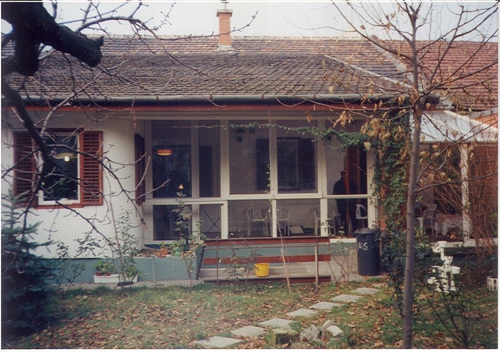 A low, pale house is seen behind a front garden with bare branches crossing the view. A wide glazed porch runs across the facade, with large panes set above a low green-gray wall. A red-brown shuttered window sits to the left, while a paved path and planting border run across the foreground. A dark wheeled bin stands near the right end of the enclosure.