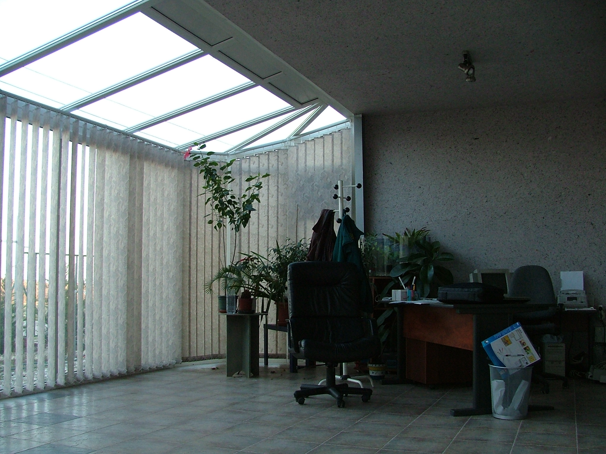 The interior room has a slanted translucent roof divided by white framing and a broad wall of vertical blinds. Several potted plants stand near a desk area with a rolling chair, smaller chairs, and office equipment. The right half is darker, with textured walls, tiled flooring, and a wastebasket holding papers.