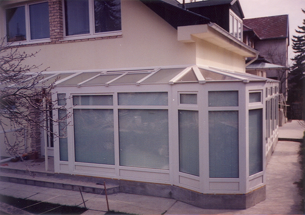 A long white-framed glazed enclosure runs along the side of a light-colored house. Its low sloping roof has multiple framed panels, and the front wall is divided into tall window sections with blinds visible behind the glass. The enclosure rests on a gray raised base beside a paved outdoor area, with bare branches at the left edge.