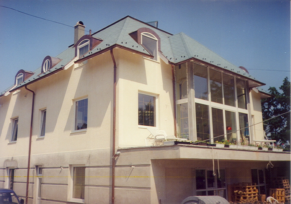 A large cream-colored multi-story house fills the frame beneath a steep pale gray roof with several dormer windows. A tall glazed enclosure with dark framing spans the right side of the house, and its panes reflect trees. White outdoor chairs stand along the enclosure, with part of a vehicle cut off at the bottom edge. Utility wires cross the sky and foreground of this low-angle exterior view.