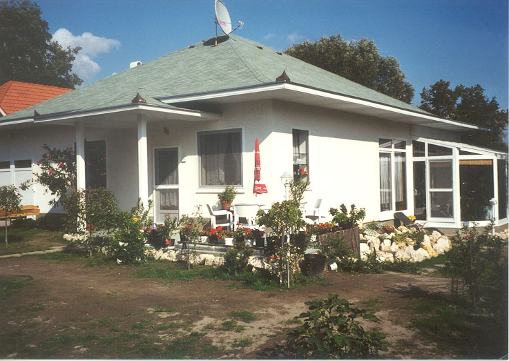 A pale single-story house has a broad gray-green hipped roof and white exterior walls. A white-framed glass enclosure extends from the right side, with chairs visible behind its panes. White chairs, a small table, and a closed red parasol sit on the front terrace among shrubs and stone edging. Trees frame the house beneath a blue sky.