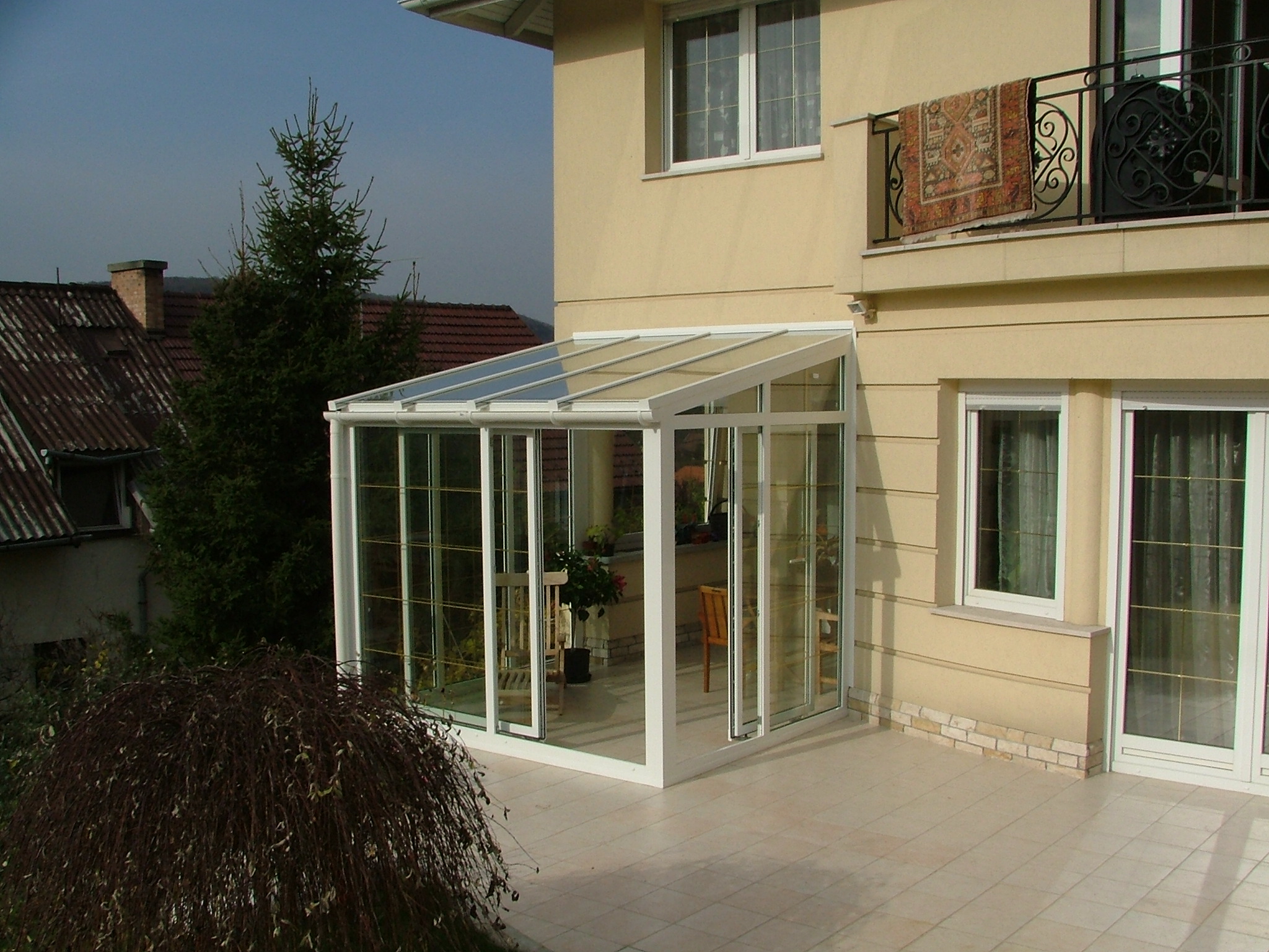 A white-framed glass conservatory with a sloping roof projects from a beige house. Narrow grid lines divide the clear wall and roof panels, through which chairs and plants are visible. Above it, a balcony has a black decorative railing and a patterned rug draped over it, while a tall tree and neighboring roof occupy the left side.