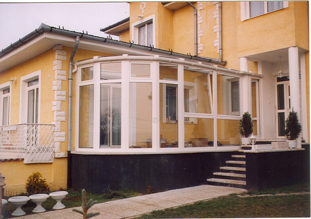 A white-framed glass enclosure runs along the front of a yellow-beige house. Its projecting window bays sit above a dark raised base, with a pale roof and white trim around the glazing. A short set of steps leads to a paved path on the right, while grass and several low white planters fill the foreground.