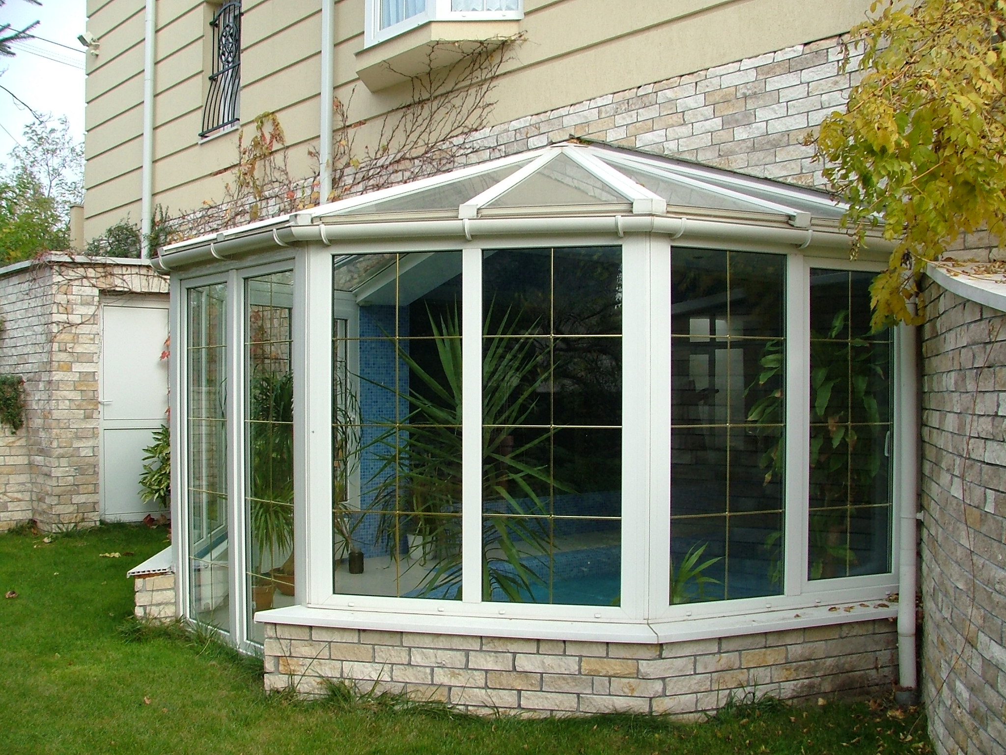 A white-framed glass conservatory extends from a beige house, with a faceted glazed roof and large gridded windows. Pale stone cladding covers the wall behind it and the base below the windows. Green plants are visible through the glass, with a lawn and leafy branches in the foreground and at the right edge.