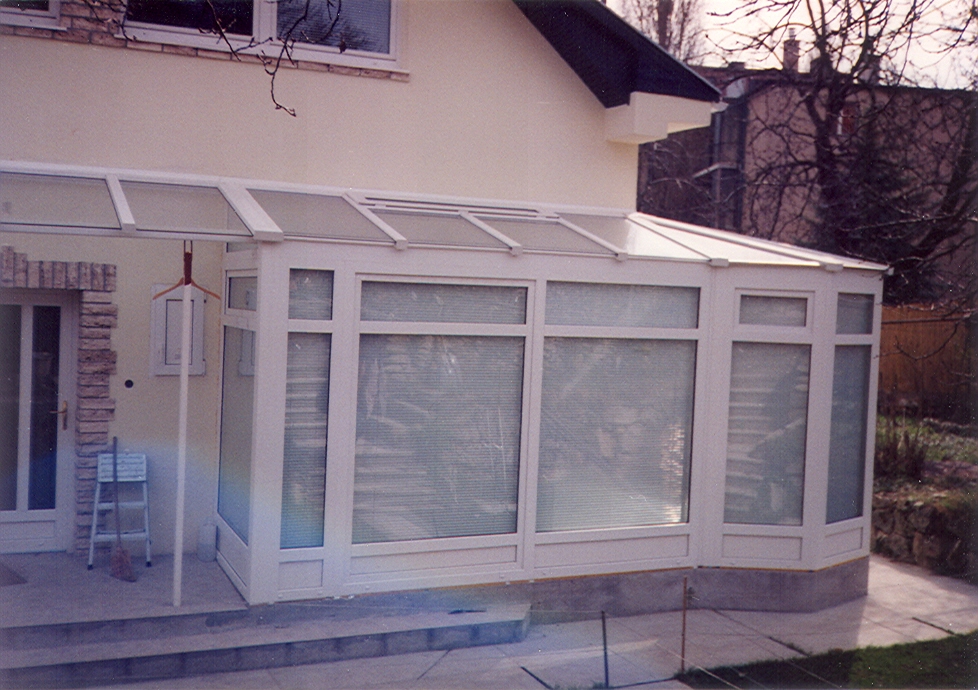 A white-framed glazed enclosure extends from a light-colored house across a paved patio. Its sloping roof and front are divided into rectangular panels, with blinds visible behind much of the glass. A glass door and a small chair sit at the left, while bare branches and a stone wall appear behind the structure on the right.