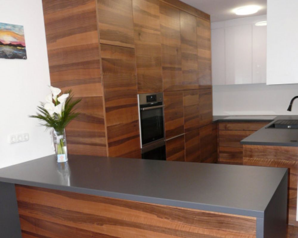 Close view of a wood-grain kitchen with dark island worktop, tall cabinets, built-in oven, and white wall cabinets.