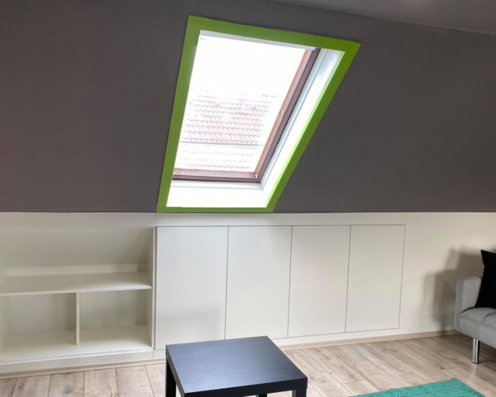 White handleless built-in attic cabinets with open shelving beneath a green-framed roof window.