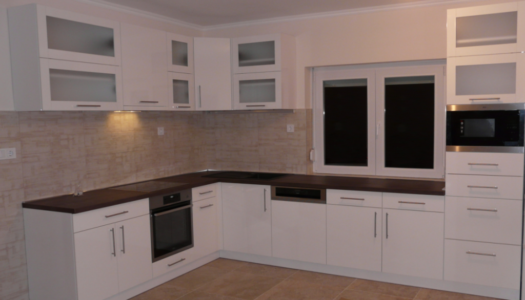 L-shaped white kitchen with dark worktops, built-in appliances, beige backsplash, and a double window.