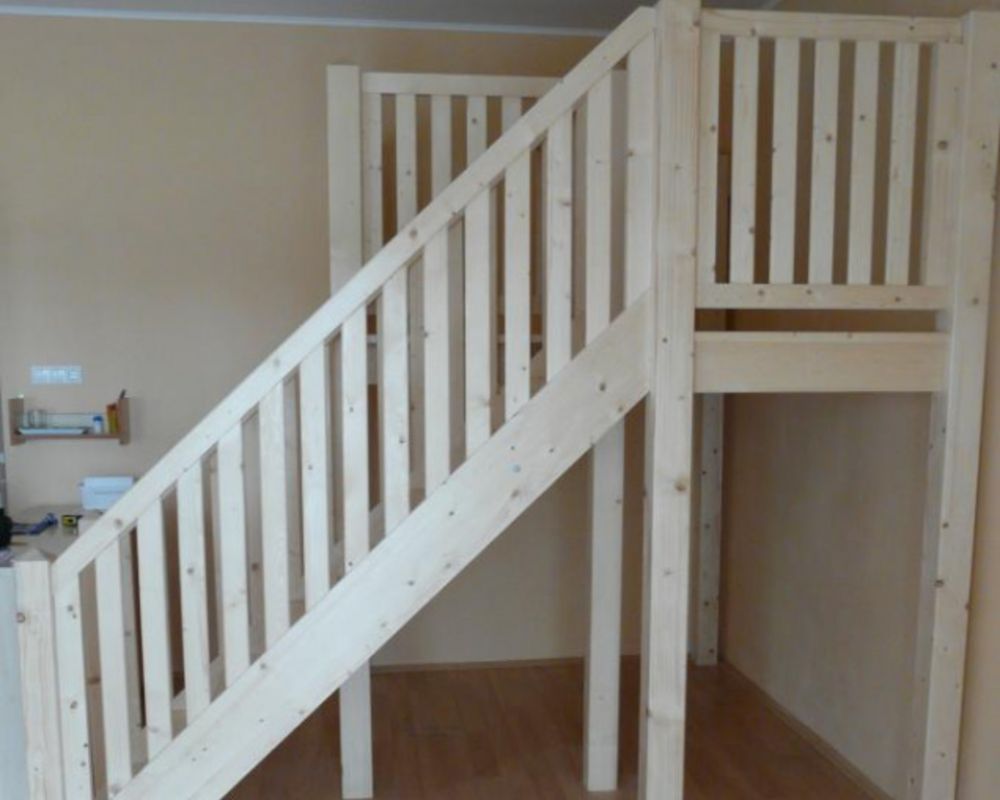 Unfinished pale wooden loft bed with a diagonal staircase and safety rails in a simple room.
