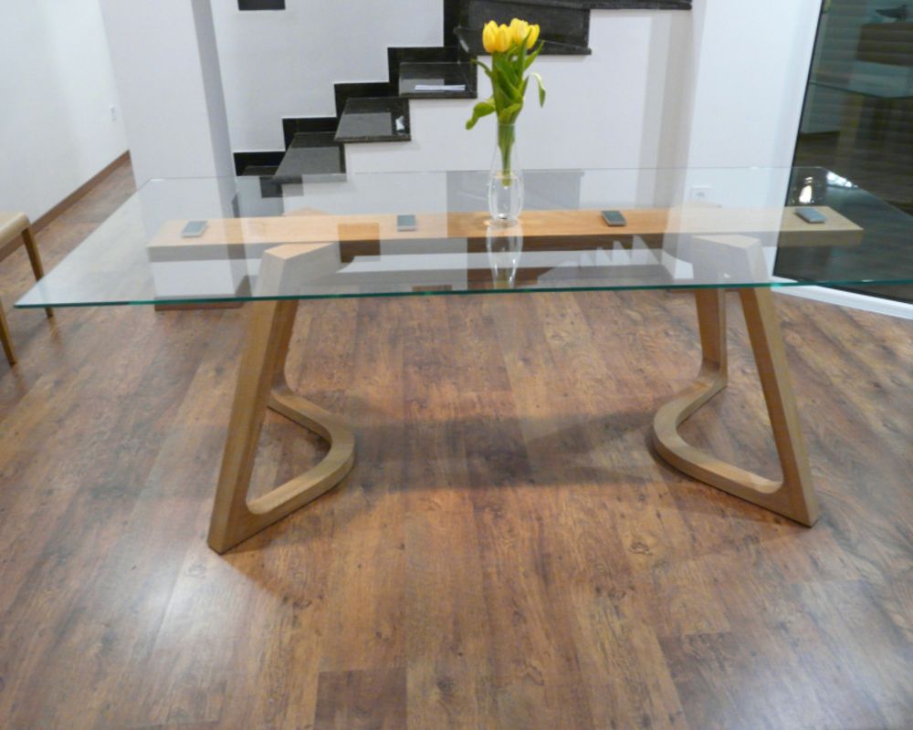 Glass-top dining table with a sculptural wooden base and yellow tulips in a bright interior.
