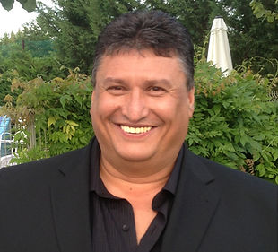 Smiling man in a black jacket posing outdoors among greenery.