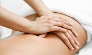Hands press gently on a person’s bare upper back during a massage. The client lies face down on a white treatment surface. The close composition uses soft neutral colors and focuses on the contact between the hands and back.