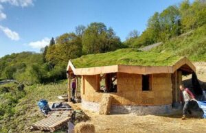 A small natural building under construction stands on a grassy hillside. Earth-colored walls support a low green roof with wide overhangs. Building materials and tools lie around the unfinished structure.