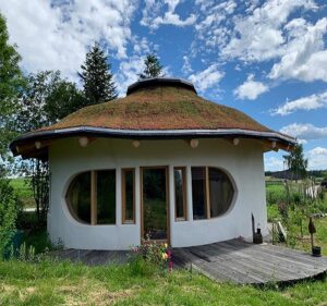 A small round white building stands beneath a broad earthen or grass-covered roof. Curved glass doors and windows face a simple wooden deck. Trees, garden plants, and a blue cloud-filled sky surround the structure.