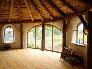 A rustic circular room has exposed timber roof framing and a light wooden floor. Tall arched windows and glass doors wrap around the curved wall. Sparse furnishings include a dark chair and small objects near the perimeter.