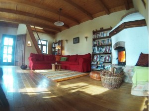 A sunlit living room has a wooden floor and a curved timber ceiling. A red sofa stands beside bookshelves and a white masonry heater. Windows and glass doors cast bright bands of light across the room.