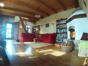 A sunlit living room has a wooden floor and a curved timber ceiling. A red sofa stands beside bookshelves and a white masonry heater. Windows and glass doors cast bright bands of light across the room.