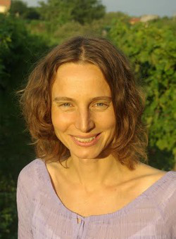A smiling woman with shoulder-length brown hair faces the camera outdoors. She wears a lavender top and stands in warm sunlight. Green foliage fills the softly focused background.