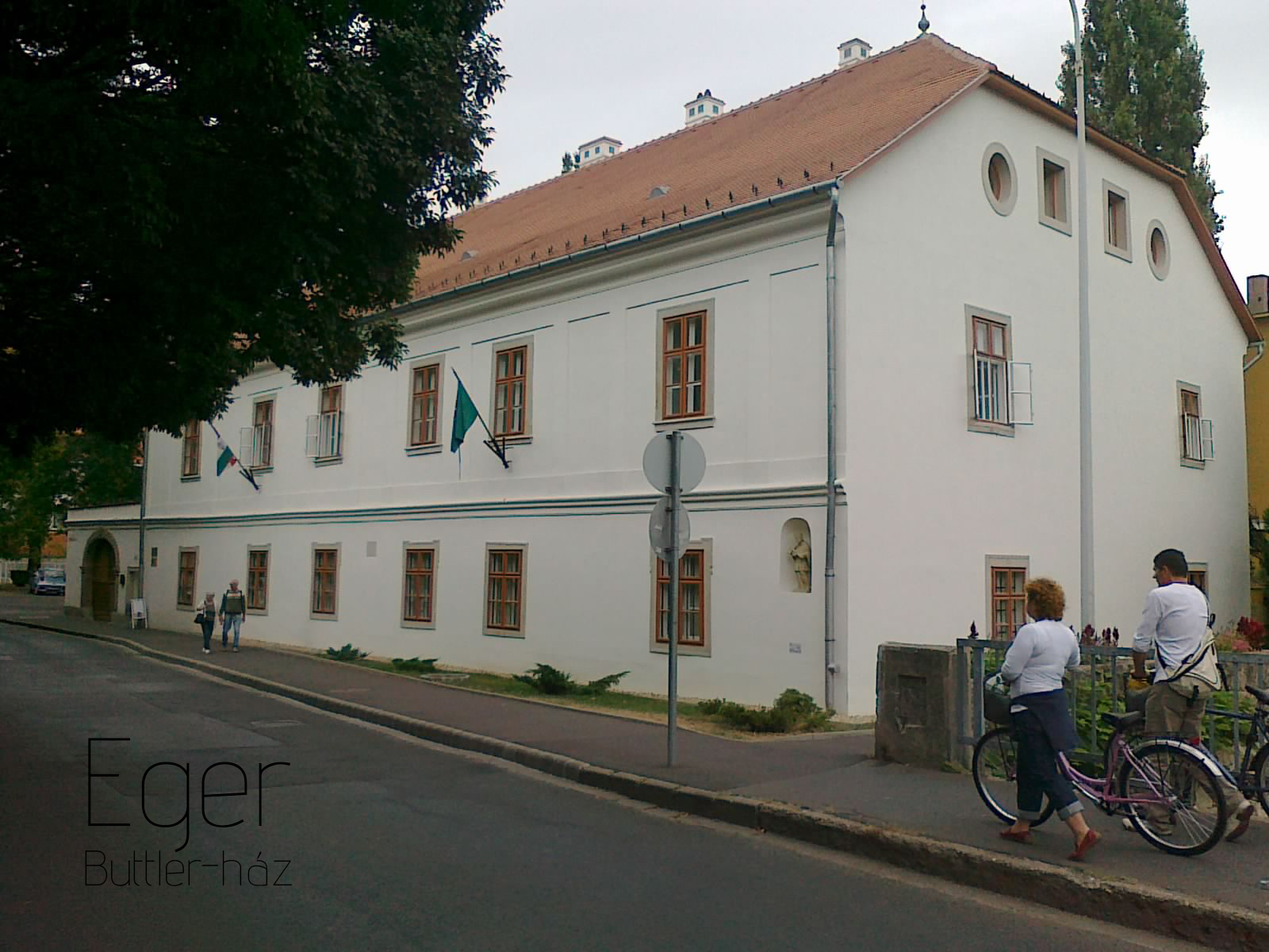 White Eger Buttler House with red roof, flags, pedestrians, and bicycles on the street.