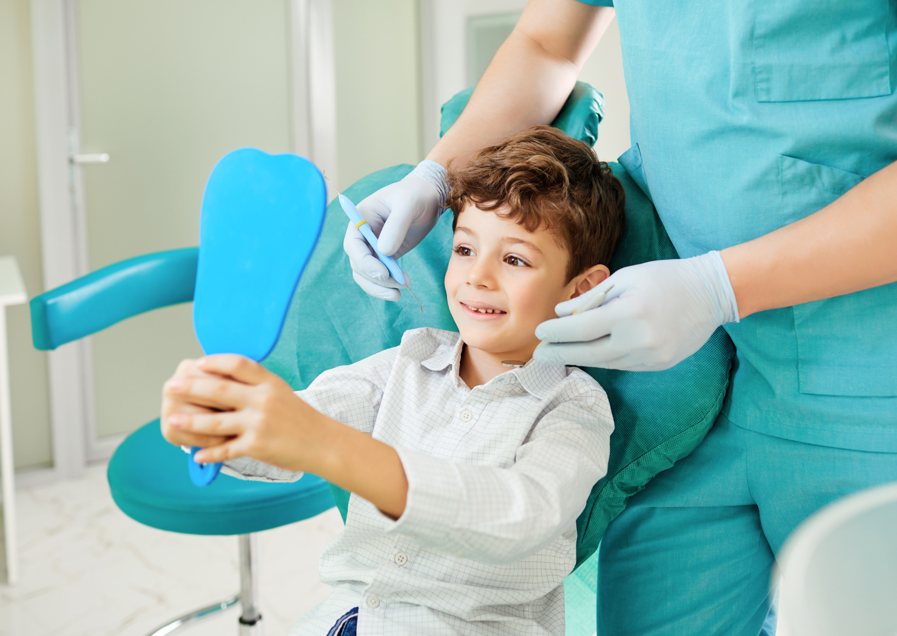 A child sits in a teal chair and holds a bright blue hand mirror while a person in teal clothing and white gloves brings small instruments near the child's face. The child looks toward the mirror, and the gloved hands frame the head from both sides. The bright room has white and aqua surfaces, with no readable text.