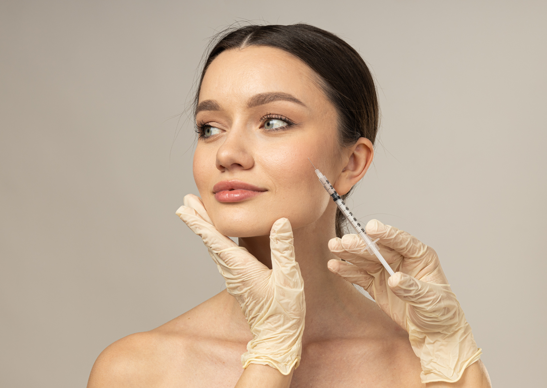 A woman faces slightly left against a plain beige background. A gloved hand supports her jaw while another gloved hand holds a syringe beside her cheek, with its needle pointed toward the skin. The crop shows her head and shoulders.