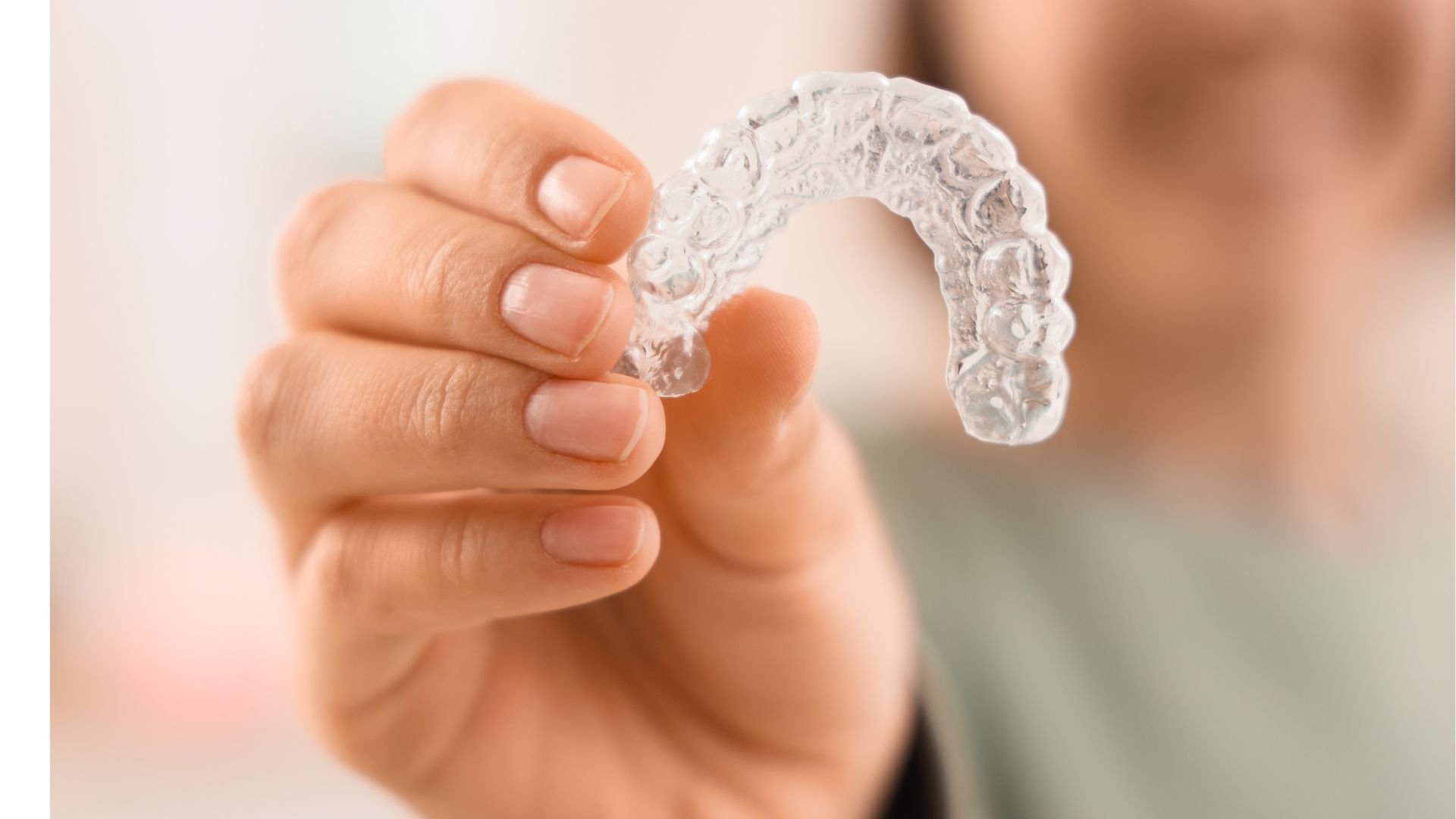 A close-up shows a hand holding a transparent dental tray shaped like an arch of teeth. The tray is lifted in front of a softly blurred face. The background is warm and pale.