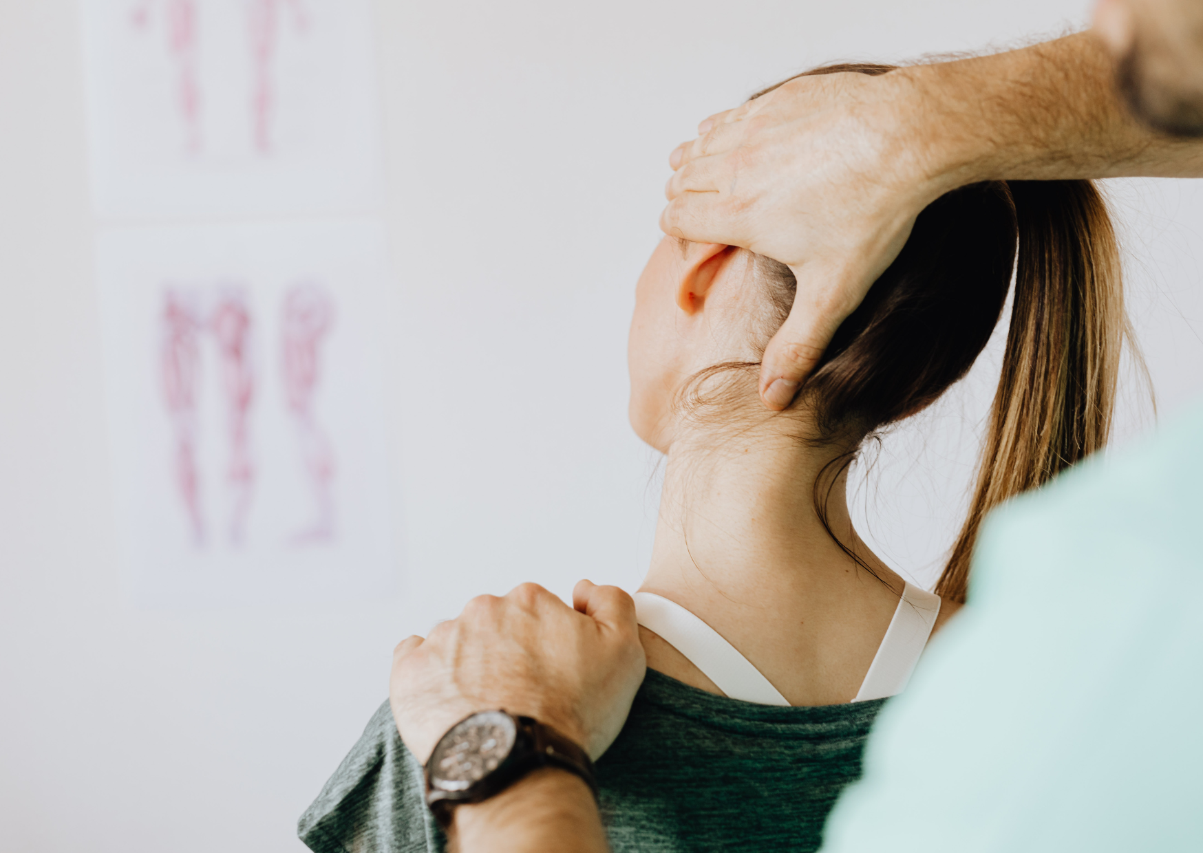A person is seen from behind with dark hair tied in a ponytail. One hand lies across the upper shoulder, and an arm comes over the head with fingers against the side of the neck below the ear. A blurred anatomical poster and pale room details appear in the background.