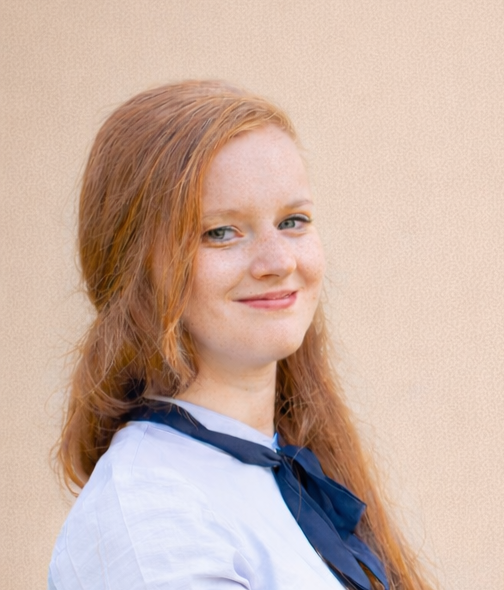 A portrait shows a woman with long copper-red hair, wearing a light blue or white top and a dark navy neck scarf. She faces the camera with a slight smile and her shoulders turned slightly to one side. A plain beige textured wall fills the background.