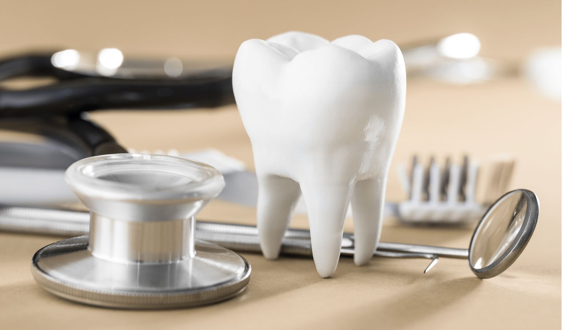 A large white tooth-shaped model stands upright on a beige tabletop beside several reflective metal instruments. A round instrument base fills the lower-left foreground, and a dental mirror leans at the lower right. Dark tubing and other tools are softly blurred behind the model, with warm cream and brown tones and no legible text.