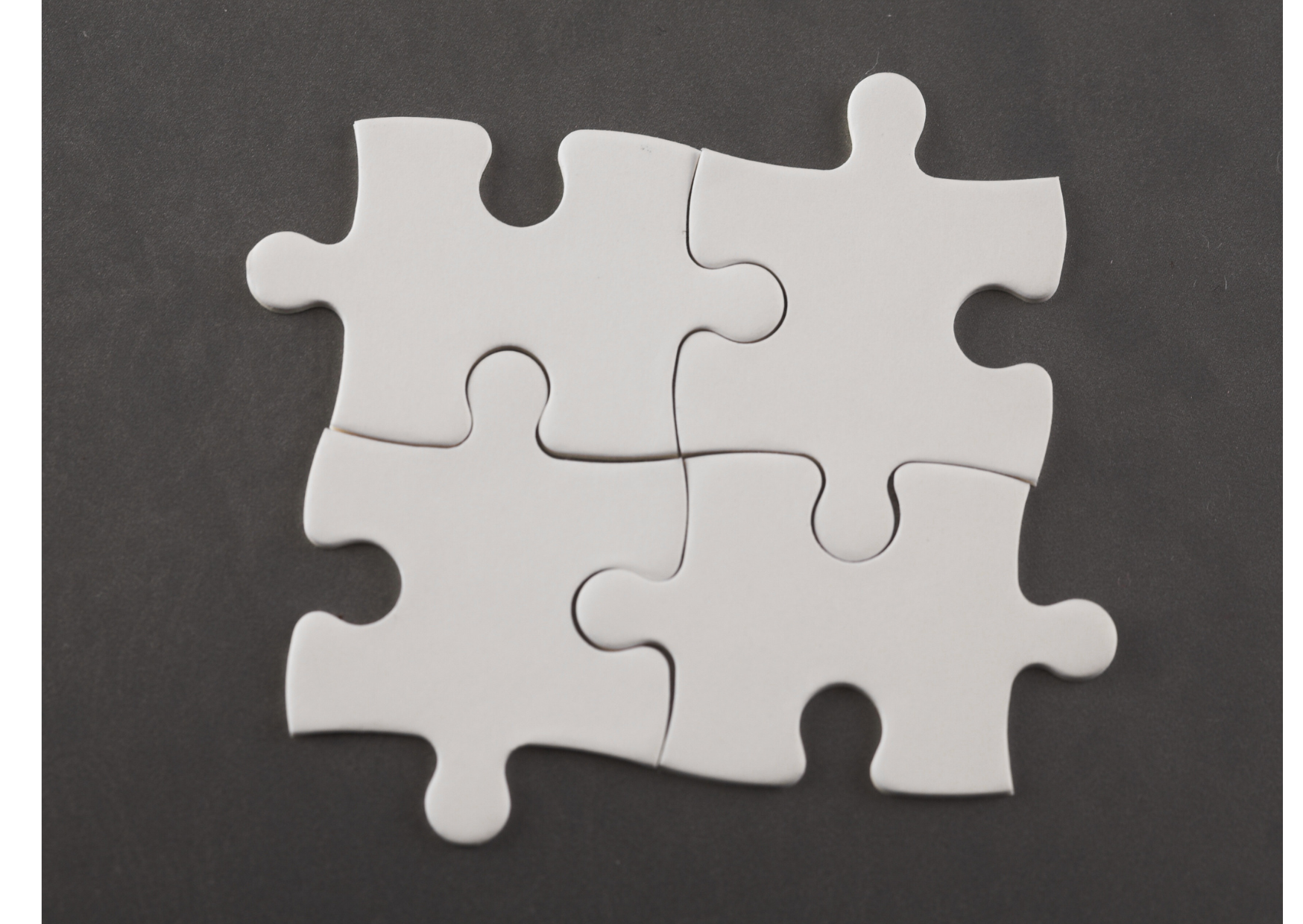 Four white jigsaw puzzle pieces lie together on a dark gray surface. Their interlocking tabs and notches form a compact square, with the piece seams visible through the center. The framing leaves a dark border around the puzzle.