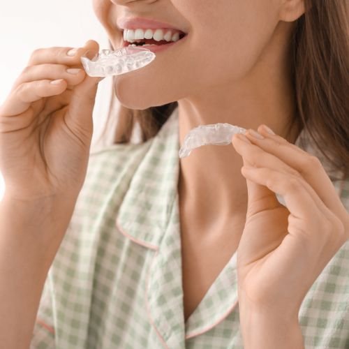A cropped view shows a woman holding two clear dental aligner trays near her mouth. One tray is close to her teeth and lips while the other is held lower in her opposite hand. A pale green checked garment and bright background fill the rest of the image.