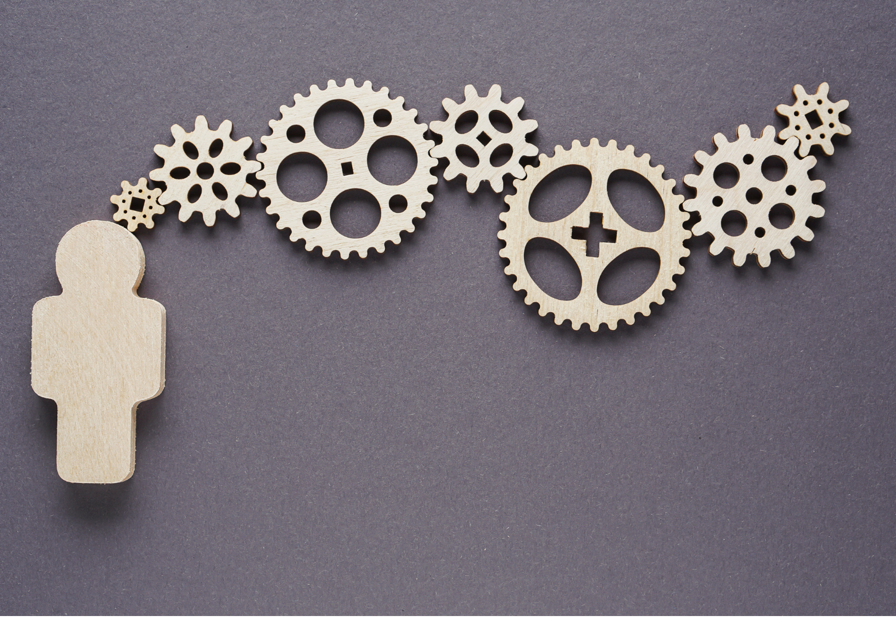 Light-colored wooden gear shapes form a row across the upper part of a dark gray surface. A larger plain wooden human-shaped cutout stands at the lower left, separated from the gears. The wood grain and dark background are visible.
