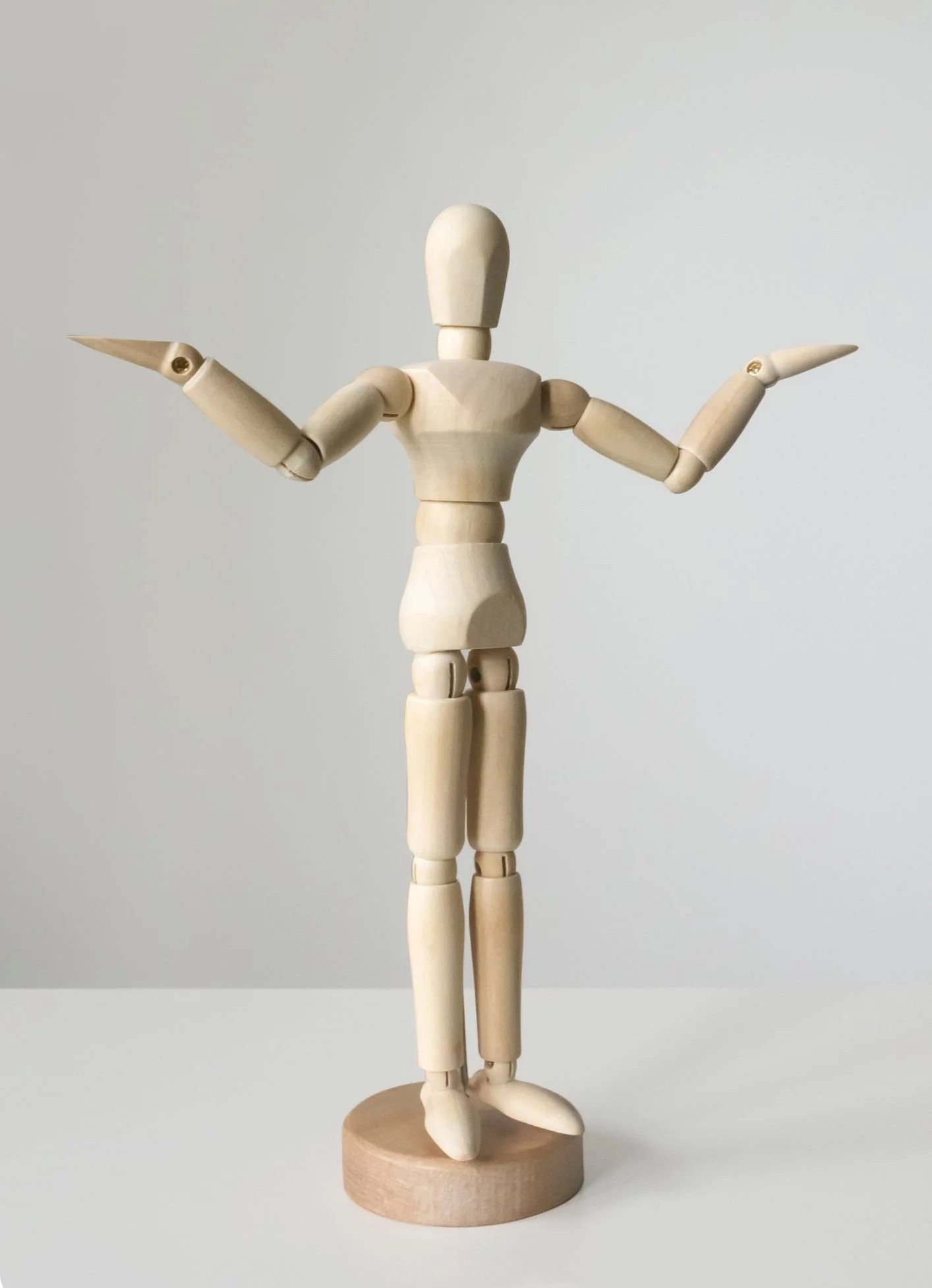 A wooden articulated mannequin stands on a round wooden base with both arms bent and raised outward. Its featureless head and segmented wooden joints are visible against a light gray background. The figure is centered in a tall portrait frame.