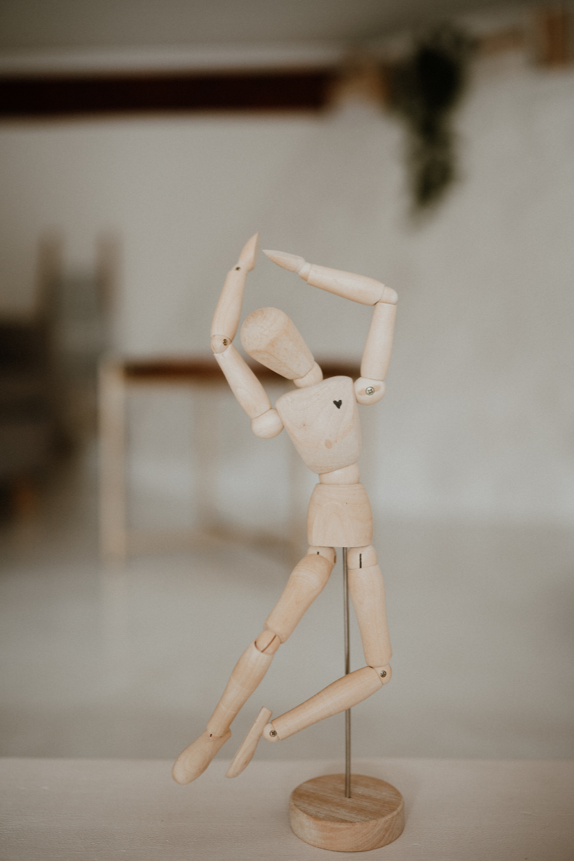 A small wooden artist's mannequin stands on a thin metal rod and round wooden base. Its arms are raised above its head, and one leg bends across the other in a posed stance. The figure sits against a softly blurred neutral background.