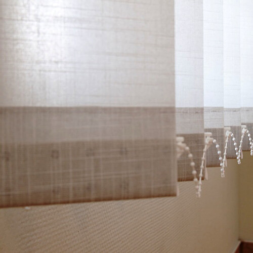 A close-up shows pale beige window-shade fabric with alternating opaque and sheer horizontal bands. The material fills nearly the whole frame, with several overlapping panels visible toward the right. White beaded pull chains hang vertically near the right edge. No people or readable text appear.