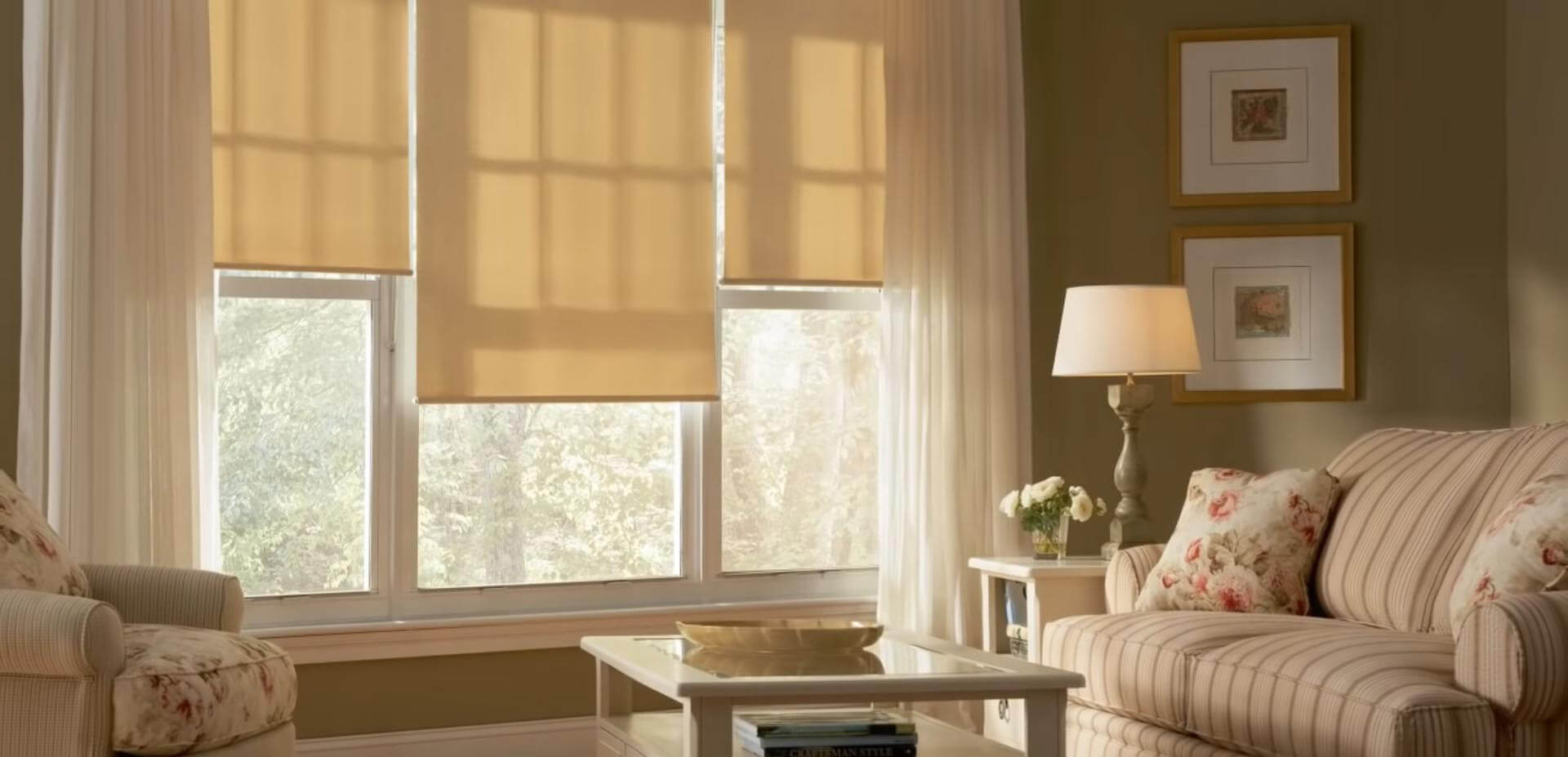 Three beige fabric roller shades hang at different heights across a bank of white-framed windows. Light passes through the fabric, leaving the window panes and leafy greenery visible below and behind them. A cream striped sofa with floral cushions fills the right side, while a matching armchair, table, lamp, and framed pictures complete the seating area. No people or readable text appear.