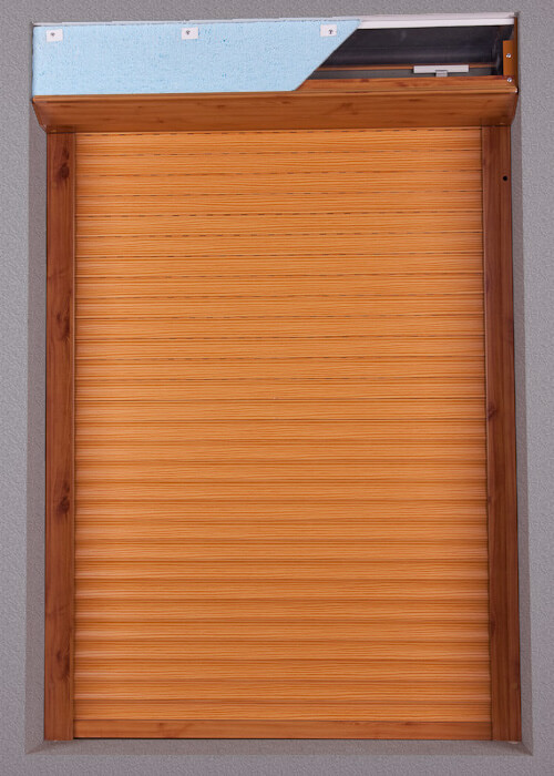 A closed shutter with narrow horizontal slats fills a rectangular frame against a gray background. The slats and side rails are brown with a fine linear grain, while the top housing has a similar finish. A light blue sheet covers much of the upper front surface, leaving a dark inset and small pale hardware exposed at the right.