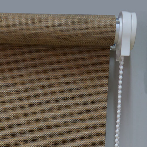 A brown mesh-like fabric shade is rolled around a horizontal tube at the top of the image. A white plastic mounting cap and beaded chain hang along the right edge, with the fabric extending downward across most of the frame. The weave forms a dense grid of small dark and light openings. A plain pale gray wall forms the background.
