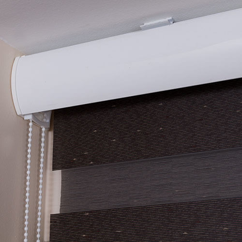 A close-up shows a dark brown striped fabric shade beneath a broad white rounded cassette mounted near the ceiling. A white beaded pull chain hangs along the left edge of the shade. The fabric has narrow alternating darker and lighter horizontal bands, with a few small speckles visible. The image is tightly cropped around the cassette, chain, and upper fabric.