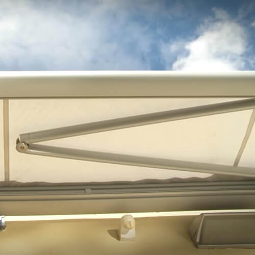 An off-white fabric canopy is extended beneath a blue sky with white clouds. A long metal support arm runs diagonally across the underside and connects to a pivot near the left side. Pale horizontal rails and a small mounting component are visible along the lower edge.