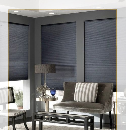 A compact interior photo shows three windows along a corner wall, each fitted with dark gray textured shades lowered nearly to the sill. A gray upholstered loveseat with a striped cushion sits in front of the center window, next to a small table and lamp. A narrow yellow rectangular border outlines much of the central image area, and parts of another chair and framed picture are cropped at the edges. The room uses gray walls and dark furniture.