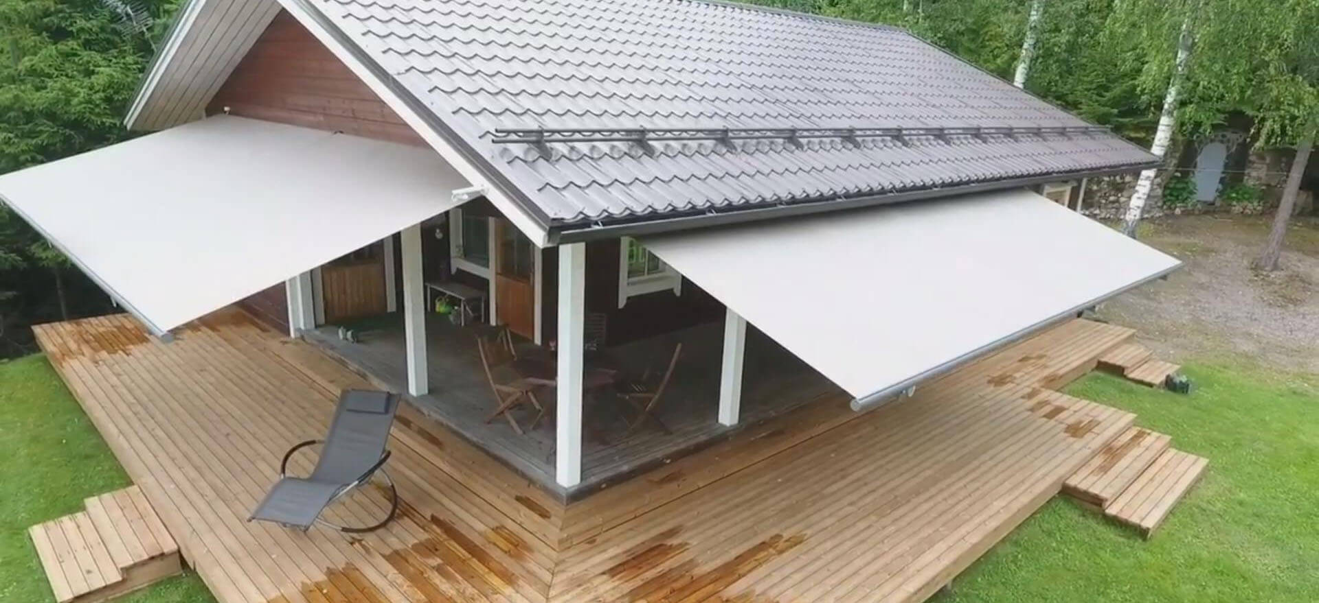 An elevated view shows a wooden house and wraparound deck bordered by lawn and trees. Two large white fabric awnings project above the deck, shading the covered outdoor area. A dark reclining lounge chair sits on the deck in front of the shaded area. No people or readable text are visible.