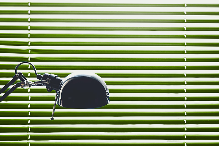 A black articulated desk lamp occupies the left foreground in front of a window covered by horizontal green-and-white slats. Narrow vertical cords or ladders run through the slats near both sides of the image. Bright daylight passes through some gaps, creating alternating bands of light and shadow. No people or readable text are visible.